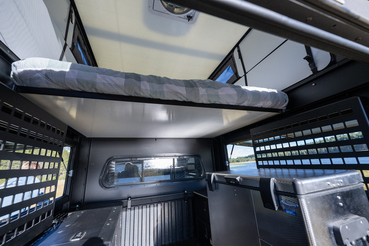 Oru Bruin camper interior with the bed platform stowed above the cabover, gear panels either side and a lake through the windows