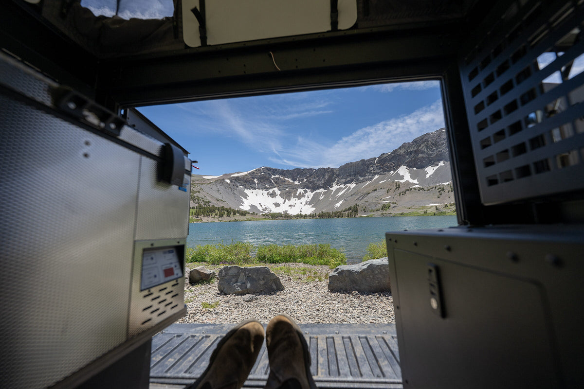 Looking out of the open rear of an Oru Bruin truck camper at an alpine lake, boots up on the tailgate