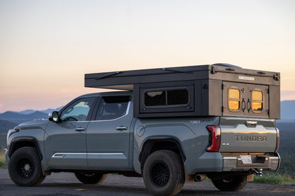 Oru Bruin XL camper on a Toyota Tundra at dusk, roof raised and the rear door windows glowing warm against the mountains