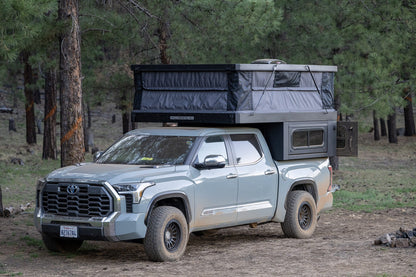Oru Bruin XL with the roof raised at a pine forest campsite, seen from the front three-quarter